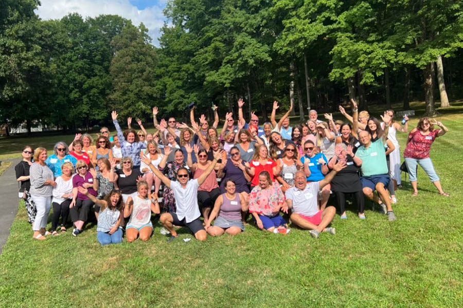About - New England Insurance Group Team Kneeling and Standing Outside on a Beautiful Day with Their Hands in the Air as They Pose for a Photo Together